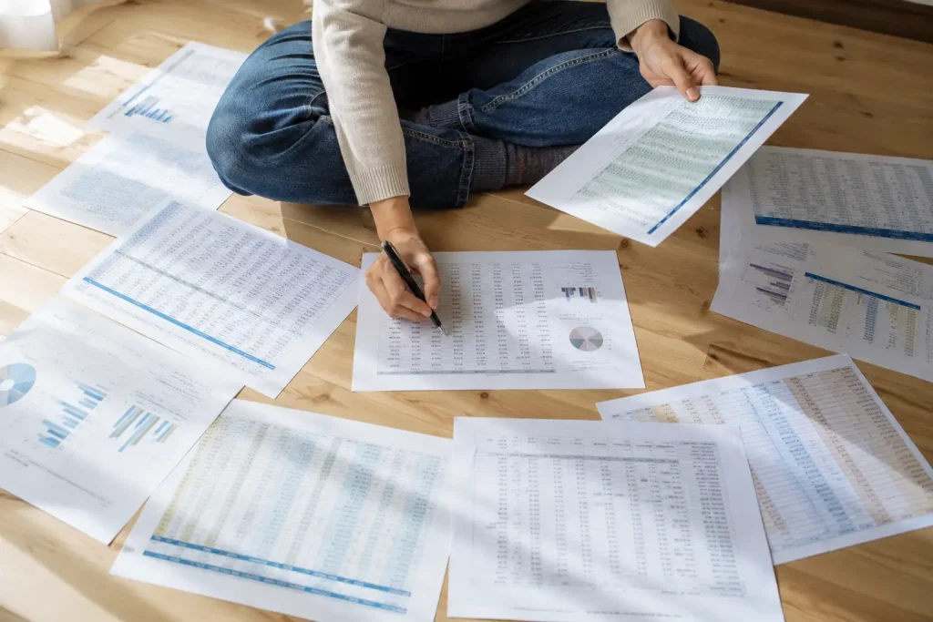 Student analizujący tabelę wyników na wydruku, siedząc na podłodze wśród papierów w jasnym pokoju