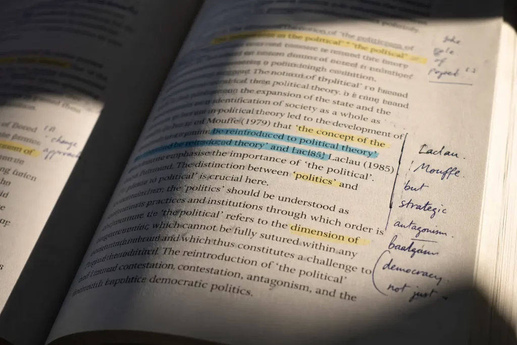 Zbliżenie na otwartą książkę naukową z zaznaczonymi fragmentami i odręcznymi notatkami na marginesach, leżącą na jasnym blacie w ostrym bocznym świetle. 