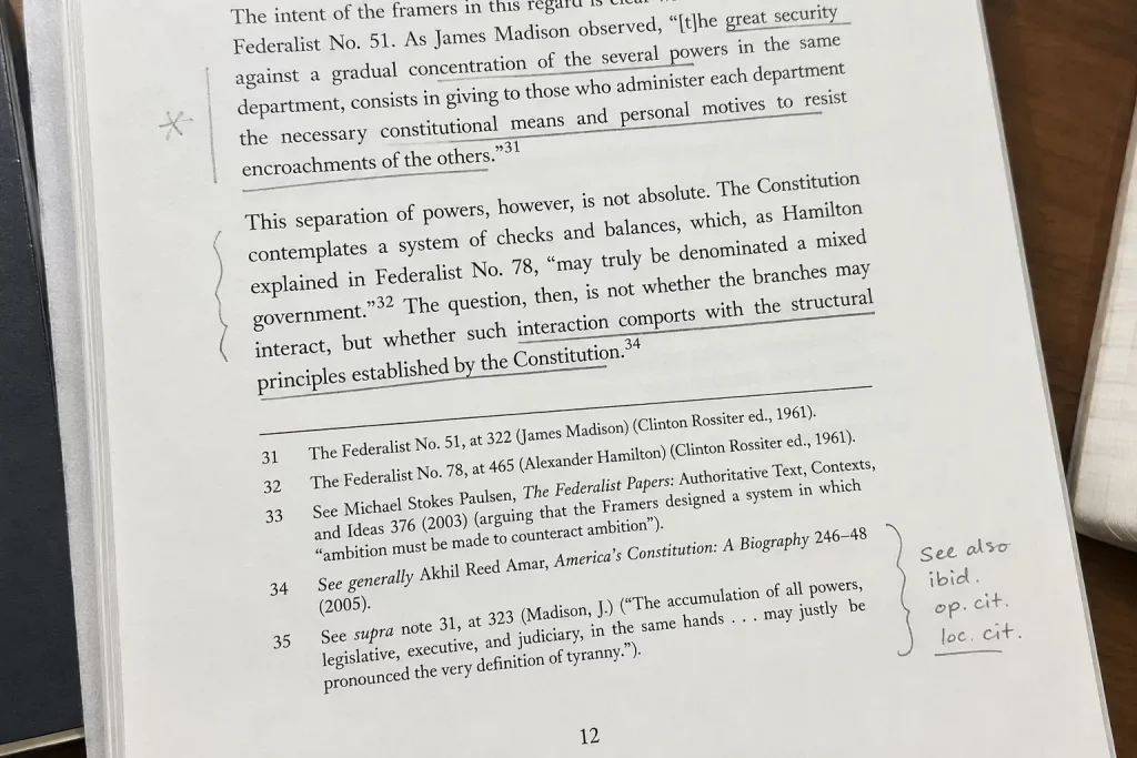  Zbliżenie na wydrukowaną stronę pracy naukowej z widocznymi skrótami ibid. i op. cit. w przypisach, podkreślenia ołówkiem, jasne biurowe oświetlenie.