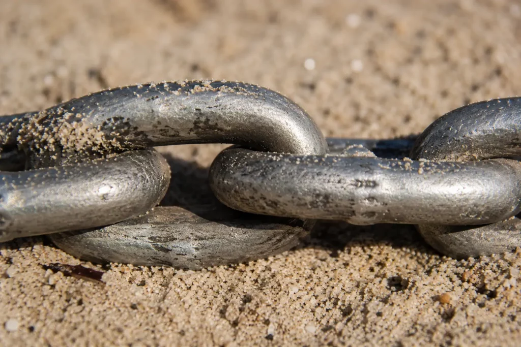 Łańcuch metalowych ogniw splecionych ze sobą leżący na jasnym piasku plaży, makro zbliżenie pokazujące dokładnie miejsce połączenia dwóch ogniw, naturalne światło słoneczne. 