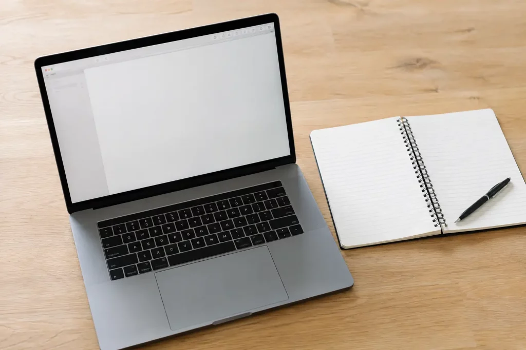 Laptop z otwartym interfejsem tekstowym na ekranie, leżący na jasnym biurku obok notatnika i długopisu