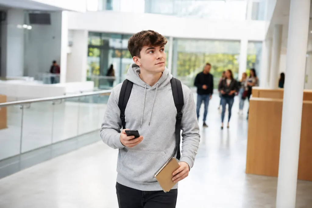 student pierwszego roku stojący w jasnym holu uczelni z planem zajęć w telefonie i notesem, w tle tablice kierunkowe i inni studenci