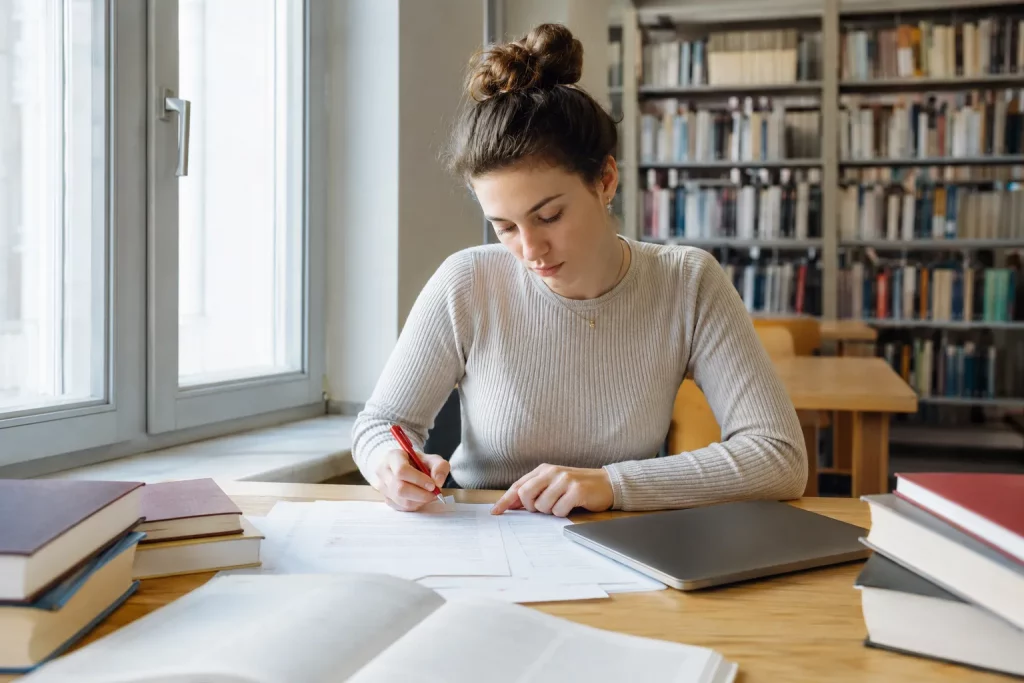 studentka siedząca w jasnej bibliotece i poprawiająca wydrukowany tekst czerwonym cienkopisem, na kartce widoczne pojedyncze znaki interpunkcyjne, regały w miękkim rozmyciu, spokojny naturalny kadr