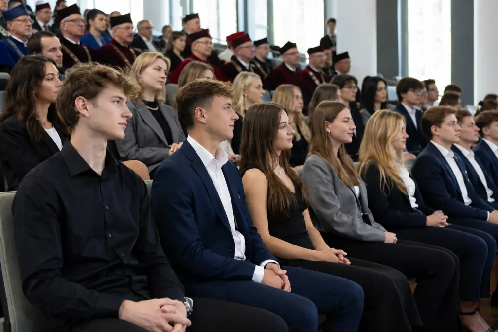 grupa nowych studentów siedząca w jasnej nowoczesnej auli podczas uroczystego rozpoczęcia roku, atmosfera oficjalna, ale swobodna